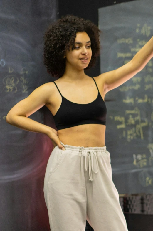 a woman wearing white pants and a black  in front of a chalk board