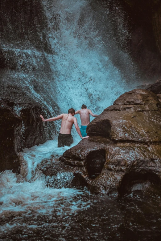 two men standing on a large rock near a waterfall
