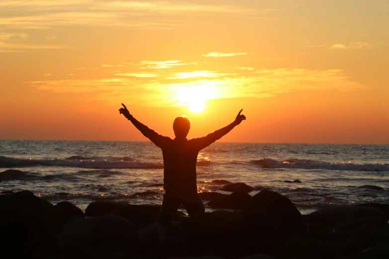 person in silhouette at sunset on rocks with arms up in front of the sun