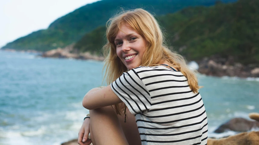 a woman sitting on the beach with a sea and rocky shoreline behind her