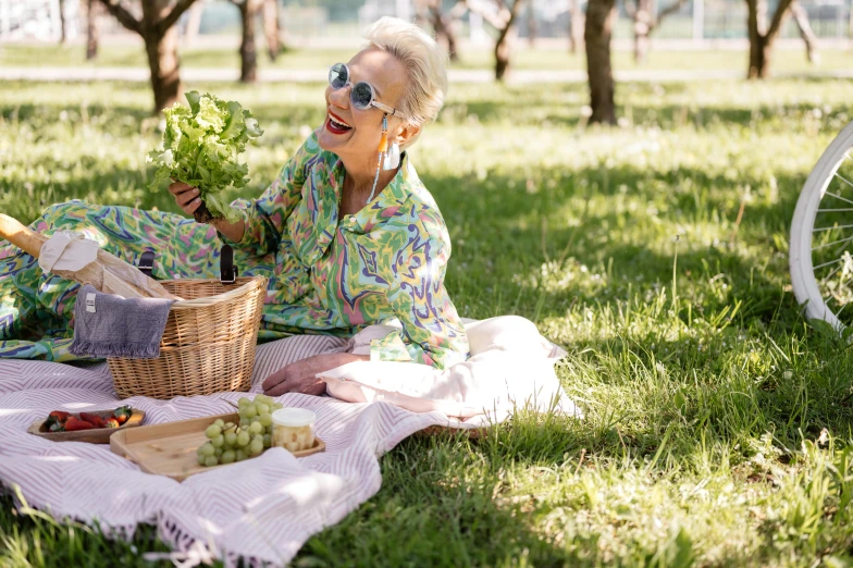 an older woman with glasses, holding a leafy vegetable, sitting on the grass