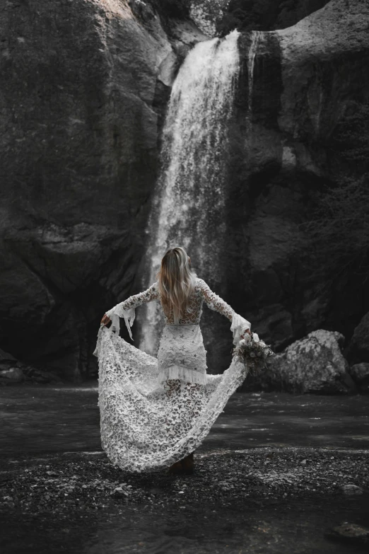 a person with a long white dress walking in front of a waterfall