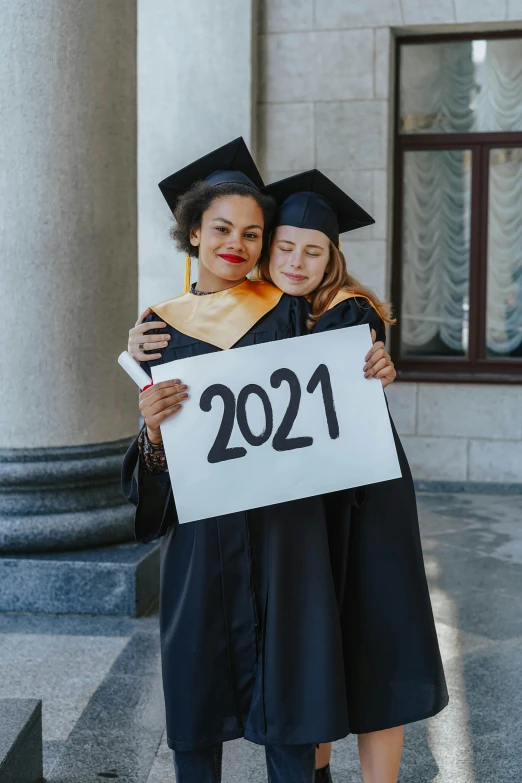 two women wearing graduation gowns and holding sign that reads 2021