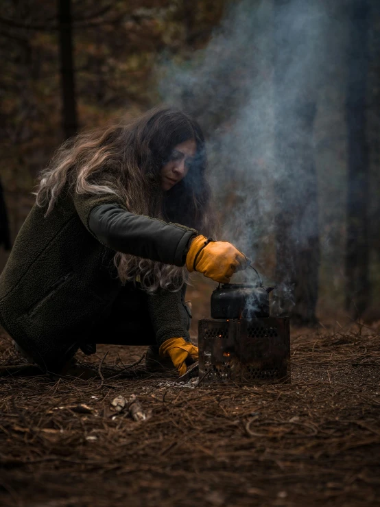 a woman wearing yellow gloves and crouching in the forest with a burning fire