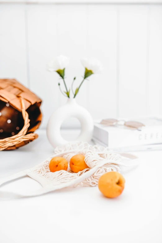 small oranges sitting on a white table near a vase and book