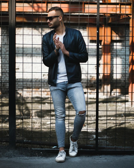 a man in ripped jeans and black jacket standing against a metal gate