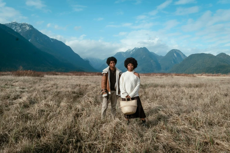 two people standing in a field with mountains and grass behind them