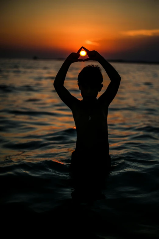 silhouette of person sitting in water at sunset, making heart symbol with hand