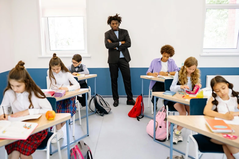 a boy stands with his arms crossed behind him as other students sit at their desks in an empty classroom