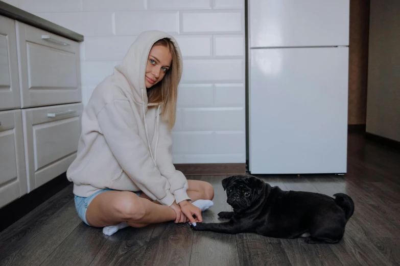 a woman in a white sweatshirt sitting on the floor petting a black pug