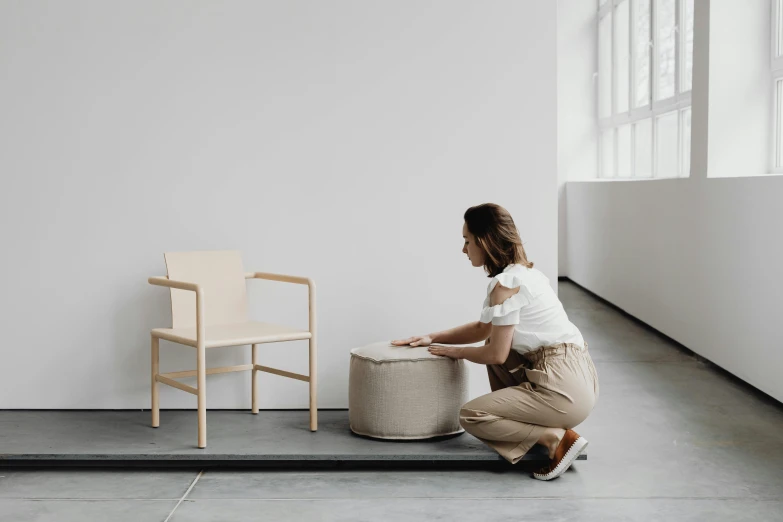 a woman crouches next to a chair in a minimal room