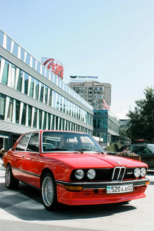 an older red bmw in a parking lot with a large building behind it