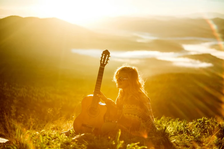 a woman sitting on a grass hill while holding a guitar