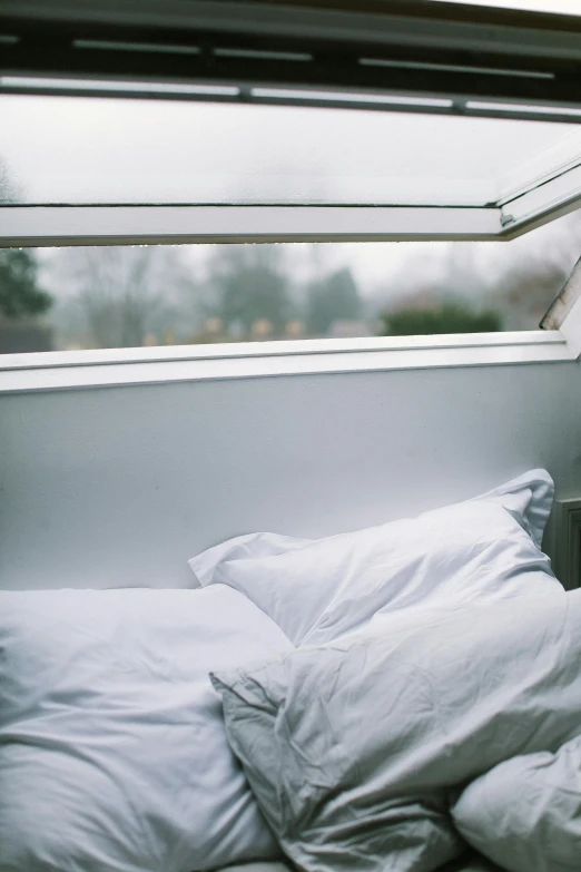 an unmade bed with white pillows and blankets, against a window