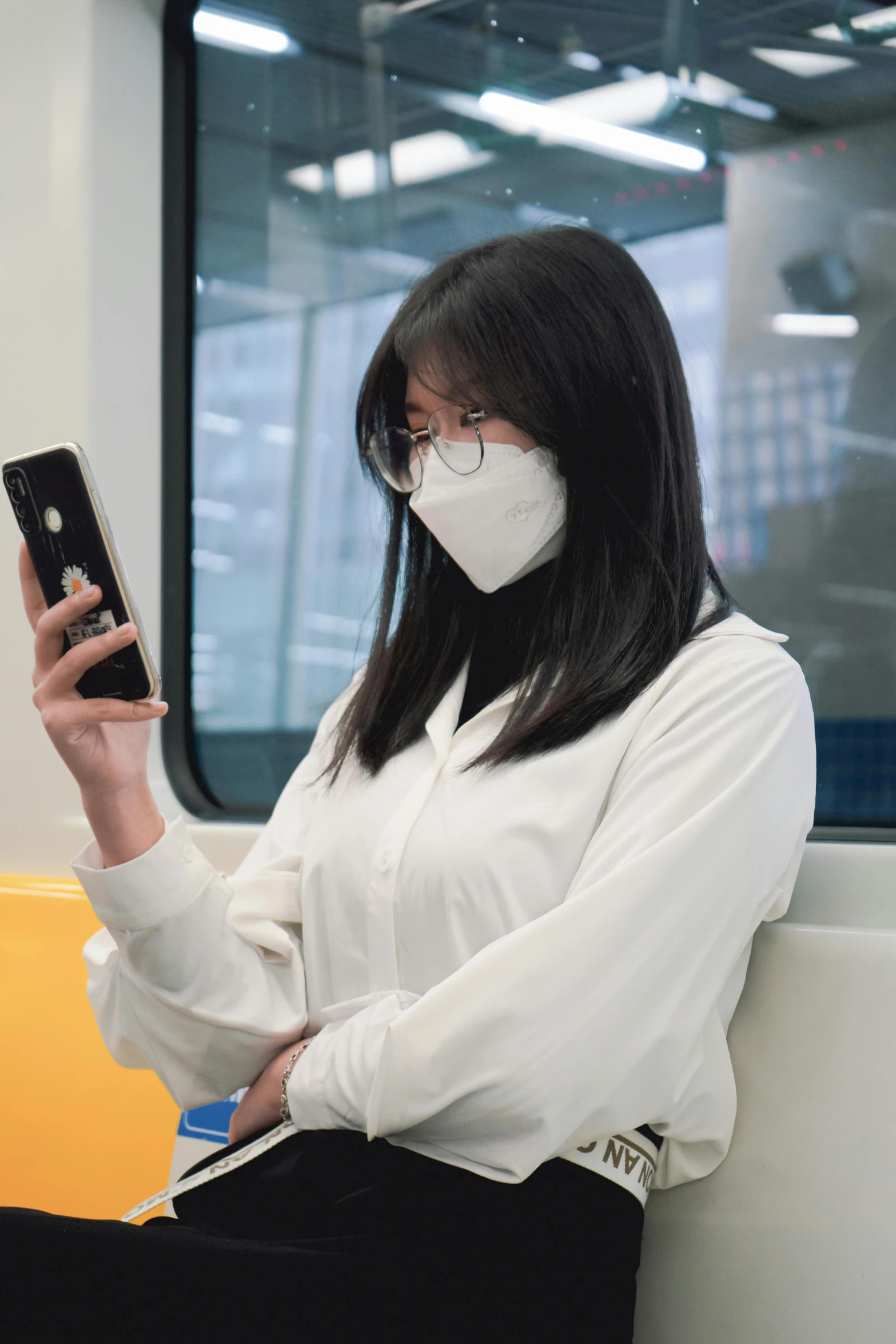 a woman wearing a white face mask looking at her cellphone while seated on the subway