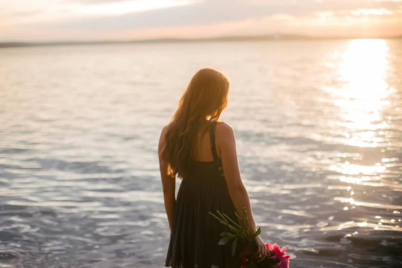 a girl in a black dress standing on the beach with a pink rose