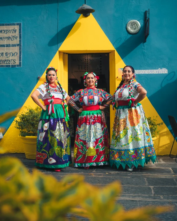 three women wearing brightly colored dresses are standing in front of the doorway of a blue building