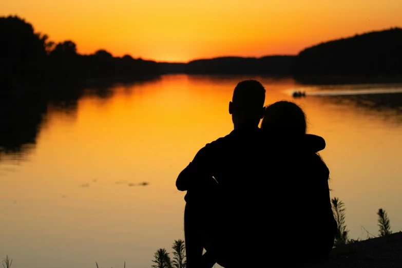 a couple is sitting by the river watching a sunset