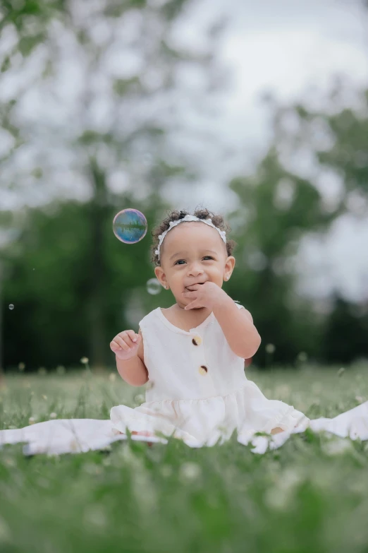 a smiling young baby girl sits on grass next to bubbles