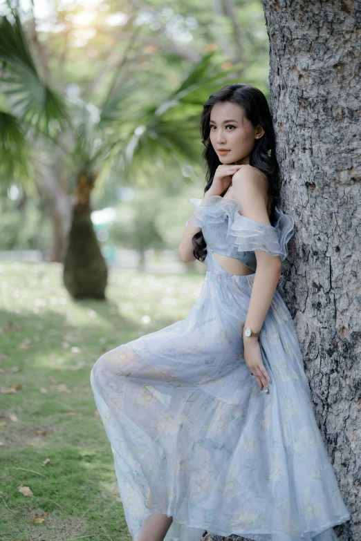 a young asian woman wearing a blue floral dress posing against a tree