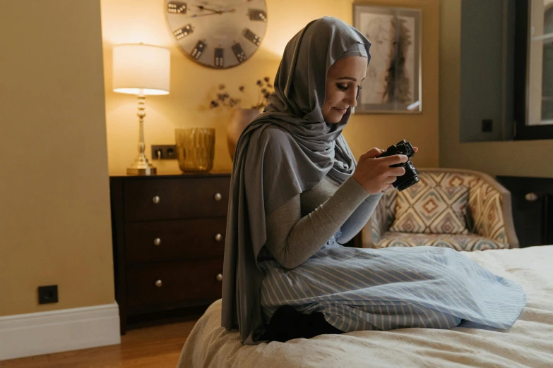 a woman in grey sweater sitting on top of bed looking at her cell phone