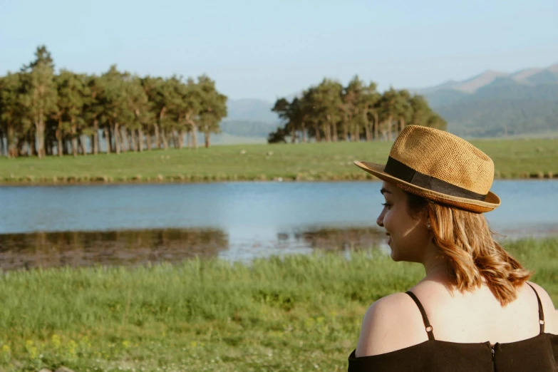 a woman standing in front of a lake with a brown hat on