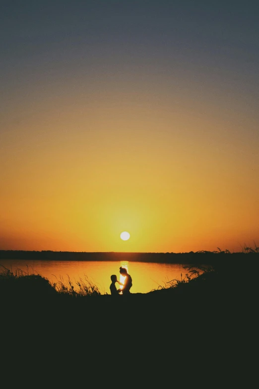 a sunset with the sun setting behind two people sitting at a lake edge