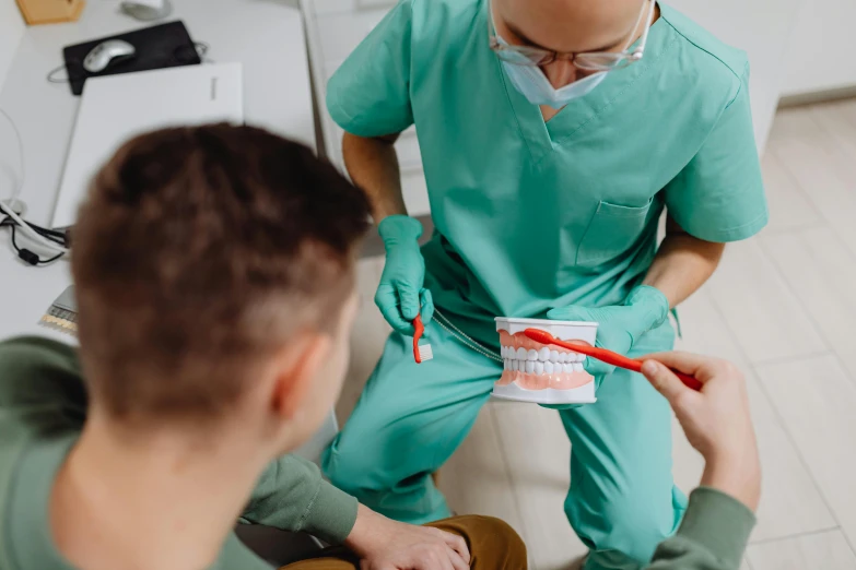 a man in scrubs sitting on a chair while another man holds a toothbrush and toothpaste