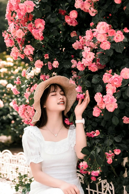 woman in a white dress and hat smiles while sitting on a bench next to pink roses