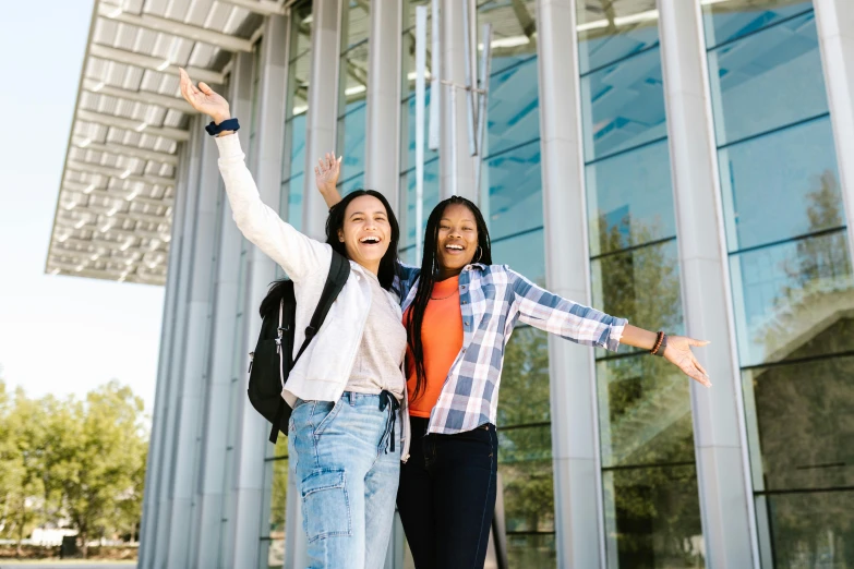 two women stand with their arms raised outside of a large glass building