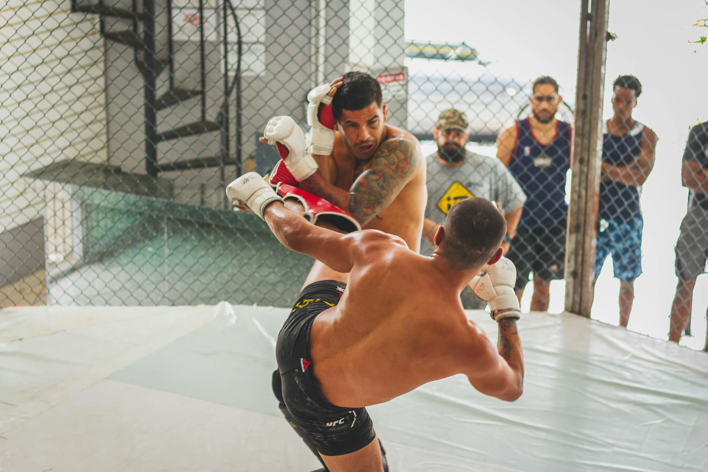 a shirtless man in black shorts and a white headgear stands in the middle of an open area as another man holds a dummy