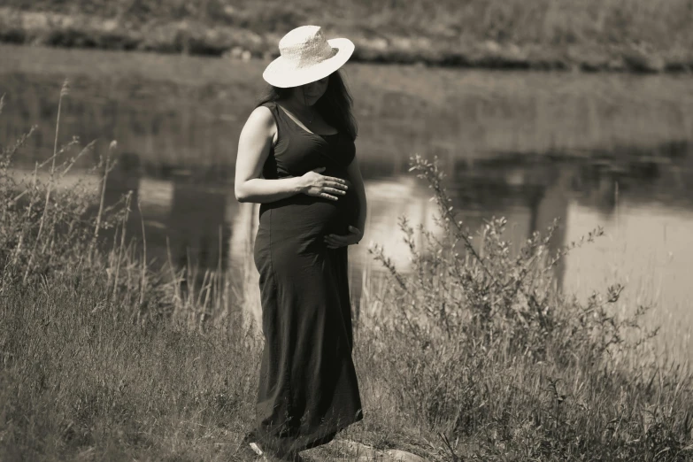 a pregnant woman in a dress and hat stands by the river