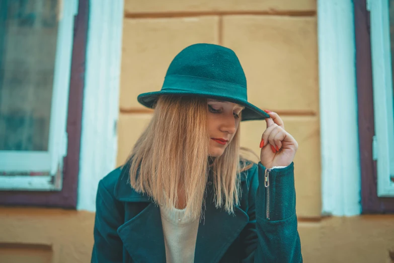 a blonde woman in a green hat poses with her hands to her face