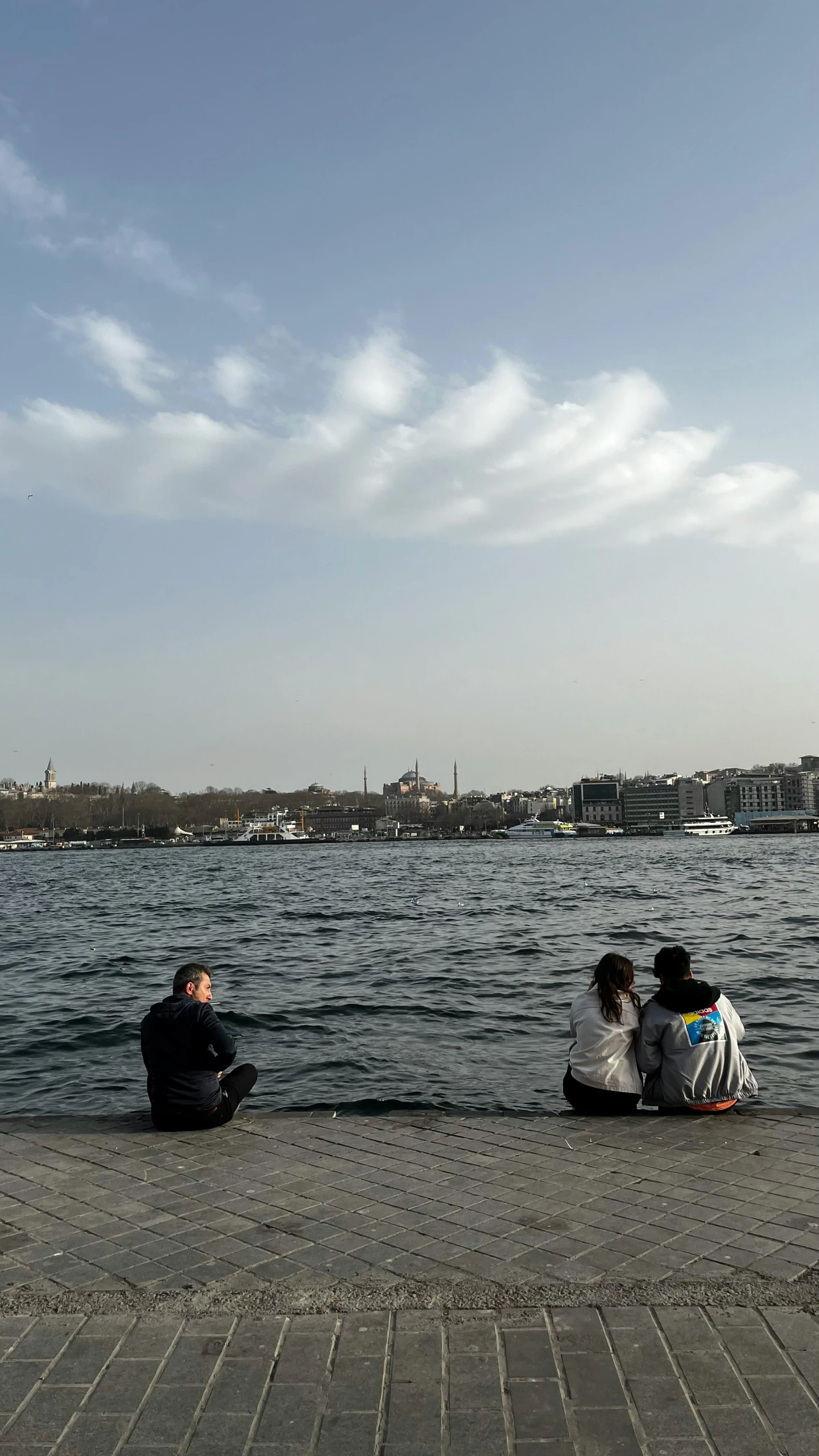 three people sit on the shore looking at a lake