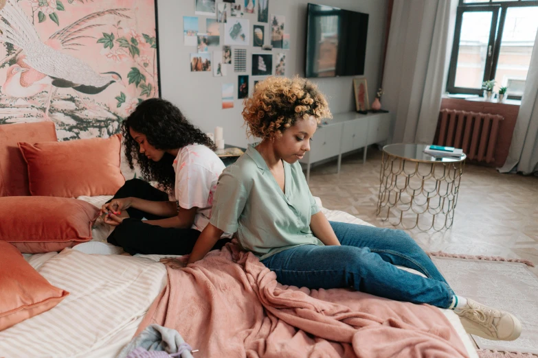 two girls sit on the edge of a bed while they look at the phone