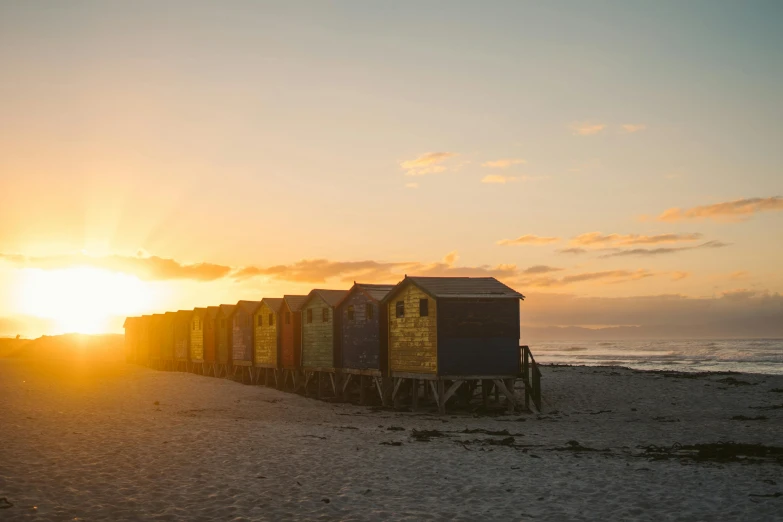 a row of beach huts in the sand with a bright sunset behind them