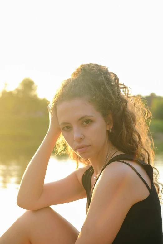 the young woman sits by the water and has a hand on her head