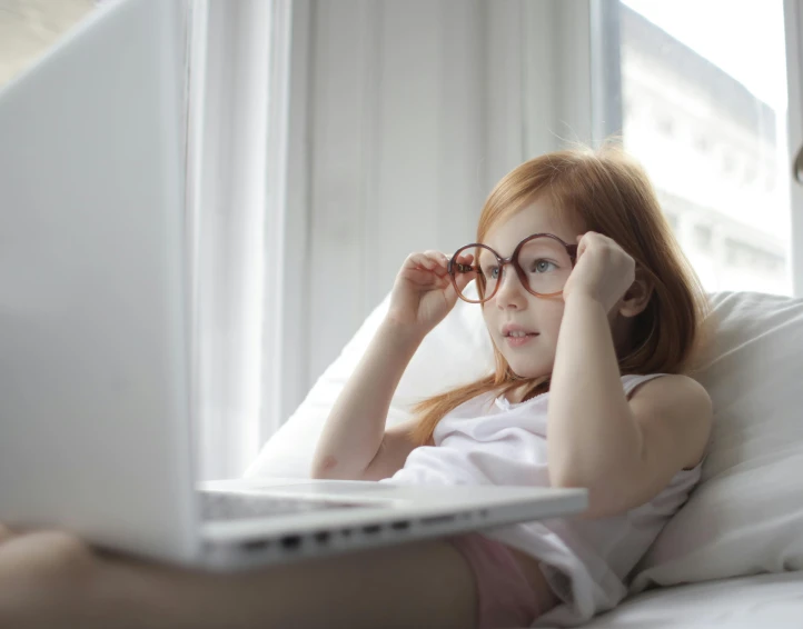a  sitting on a bed holding onto her glasses looking at a laptop