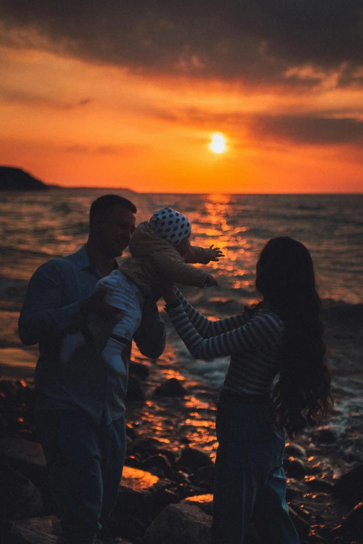 people holding the teddy bear on the shore of the ocean as a sunset looms behind them