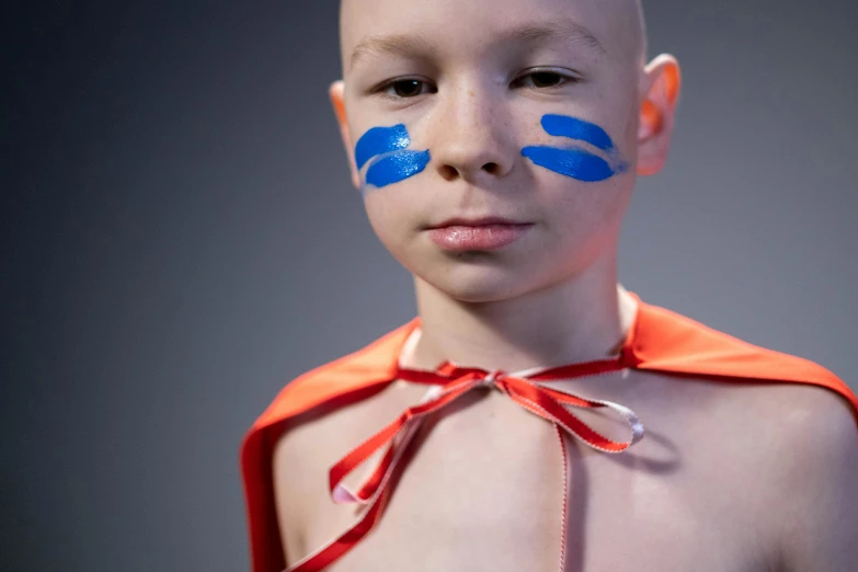 a child with blue painted on his face, and in the orange outfit, is standing next to grey background