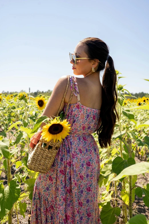 a woman wearing sunglasses, a dress and sunflowers stands in a field