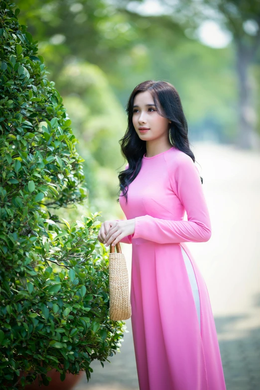 young woman dressed in pink with her hands on her waist standing in front of a tree and bush