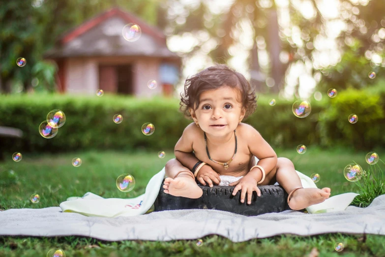 a little girl sitting on the grass in front of a blanket in the background and bubbles flying off her head