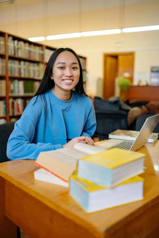 a woman smiles at the camera as she sits at her liry desk, with several books stacked on her laptop