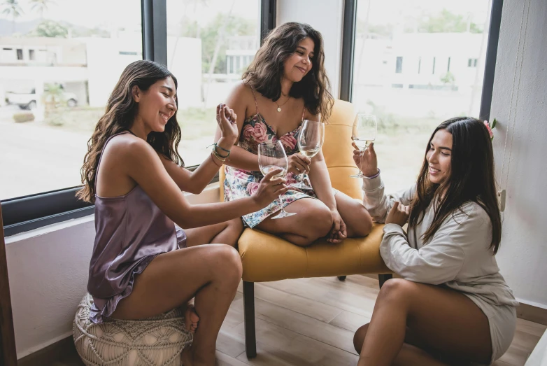 three beautiful women sit on yellow chair while one holds her wine glass