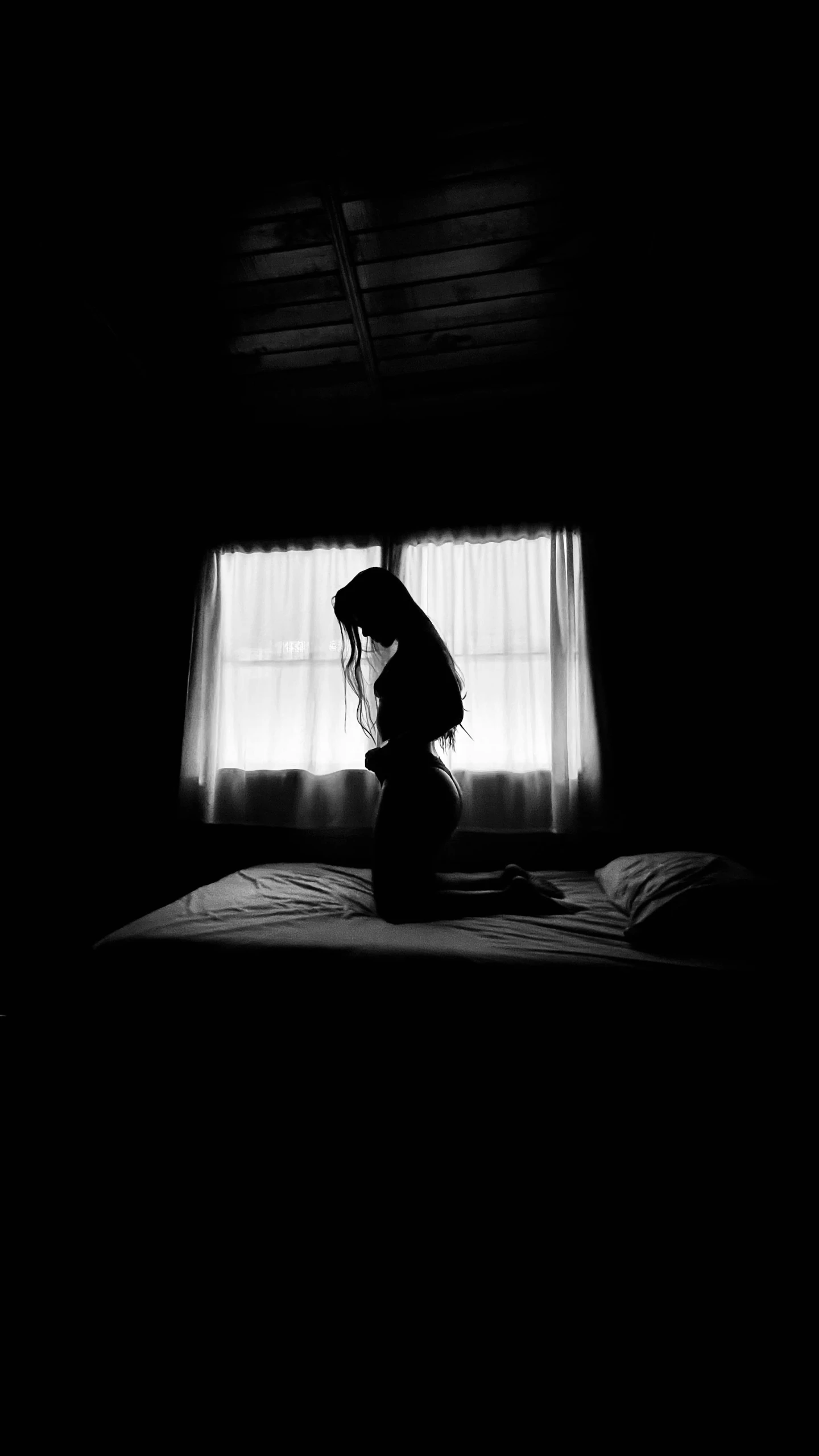 a dark room with white curtains, a woman sitting on a bed