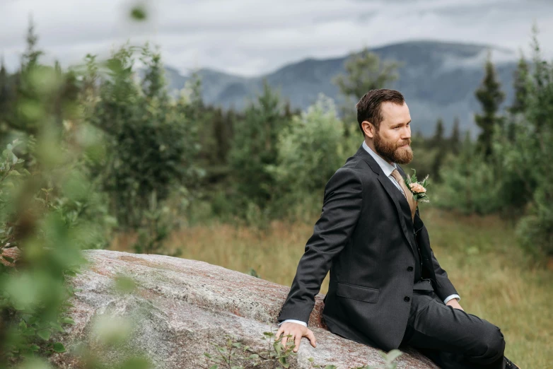 a man in a suit and tie sitting on a rock in the middle of trees