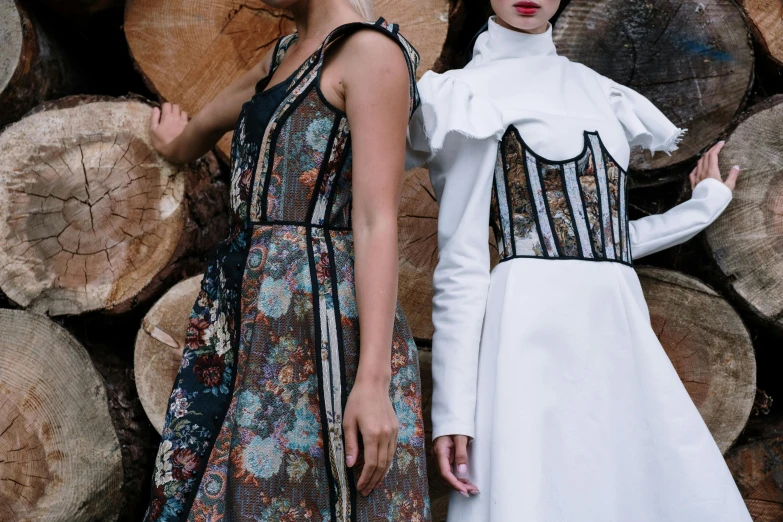 two woman in dresses are standing in front of a stack of wood