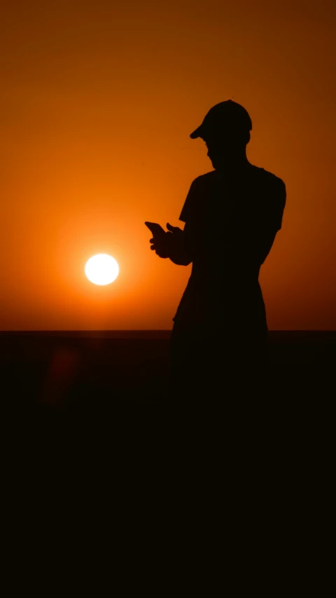 a silhouetted person wearing a hat holding his hands in his pocket while looking at the sun
