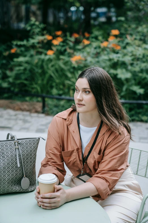 a girl sitting at a table in a park with a cup of coffee